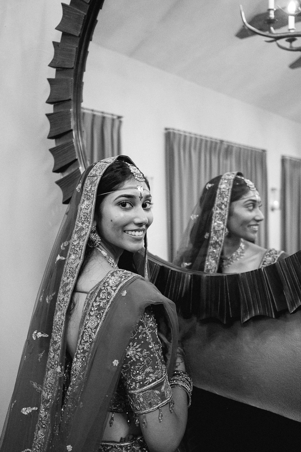A woman dressed in traditional Indian attire smiling at the camera, with her reflection visible in a decorative mirror.