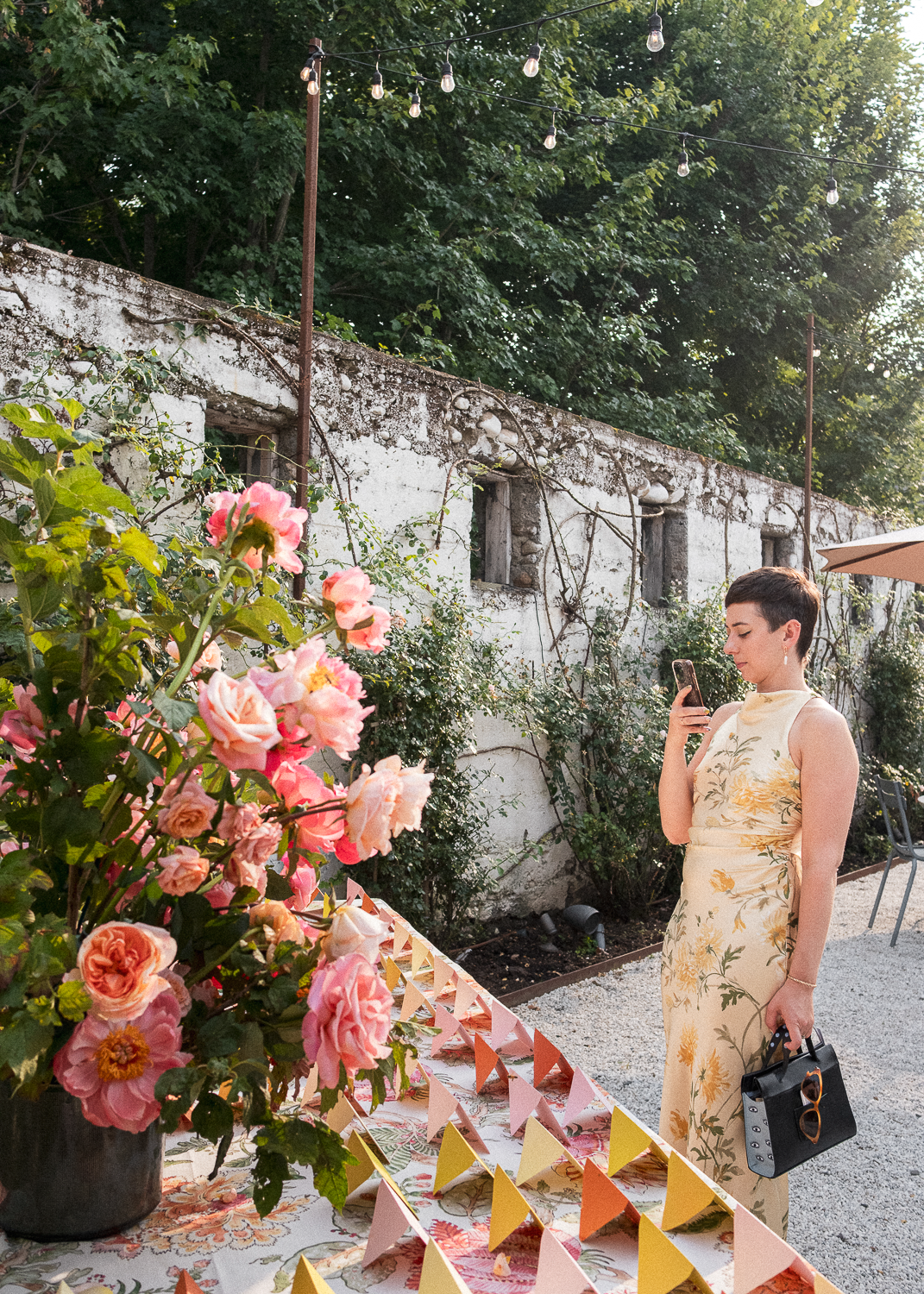 A woman in a sleeveless yellow dress with floral patterns standing outdoors, looking at her phone. She holds a black handbag and a pair of sunglasses. In front of her on a table are pink and peach flowers and colorful triangular paper decorations. Th