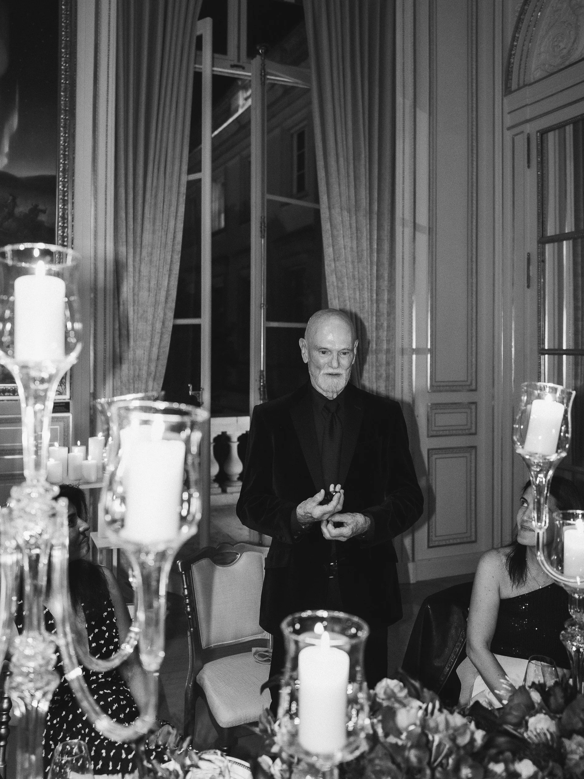 A man in a tuxedo speaking at a formal dinner event, with guests seated around a table decorated with candles and floral arrangements, in an elegant room with large windows and ornate walls.