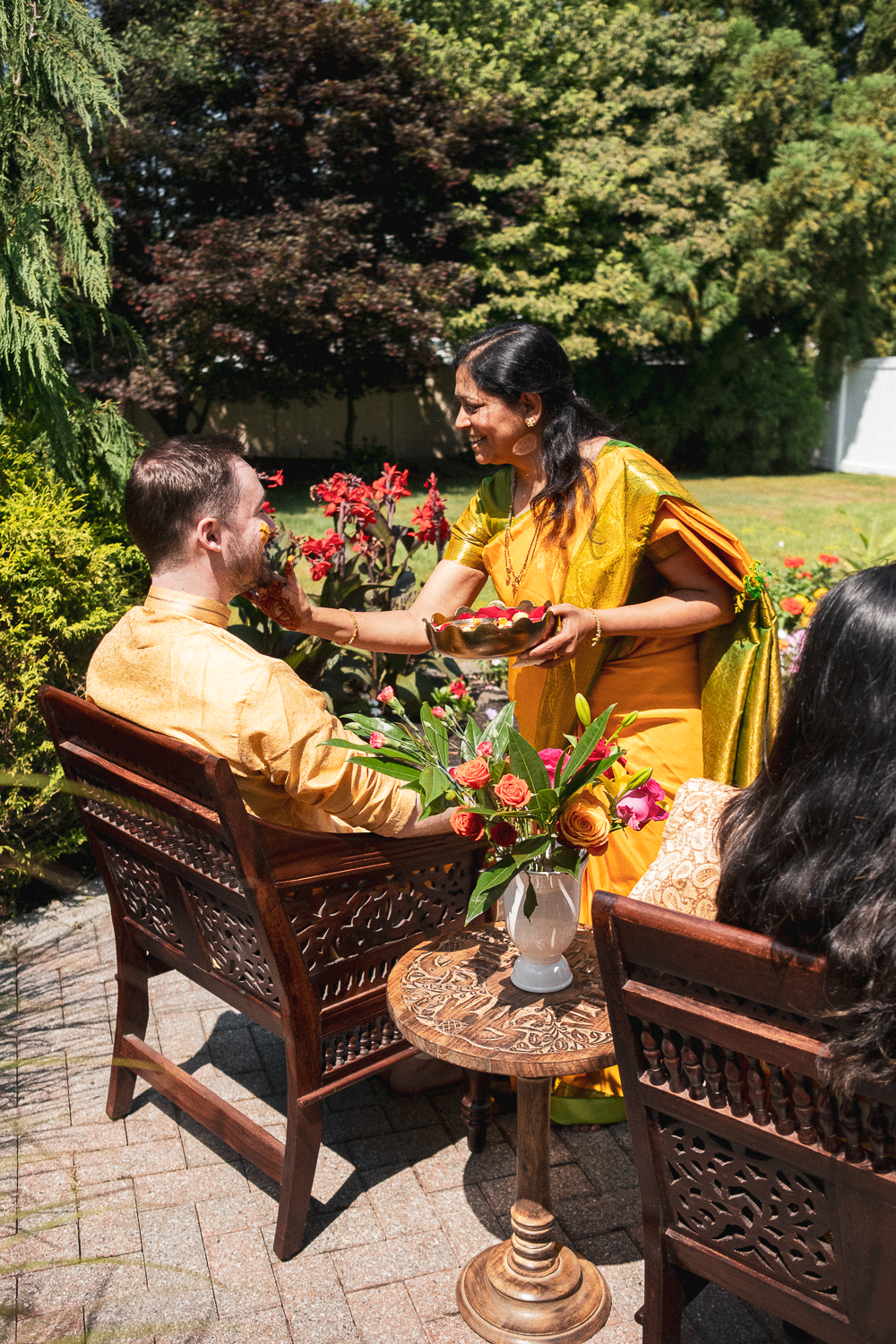 A woman dressed in traditional Indian attire offers a flower to a man sitting in a garden during a celebration, with a vase of roses on the table between them.