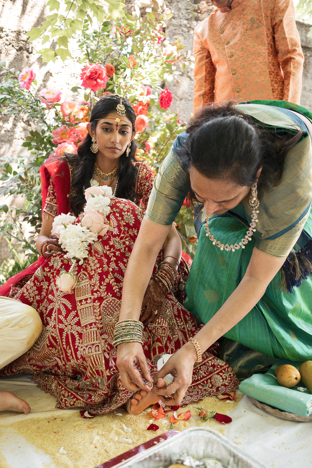 Indian bride dressed in red and gold wedding attire, seated with floral arrangements, while a woman in green performs a traditional ritual during a wedding ceremony outdoors.