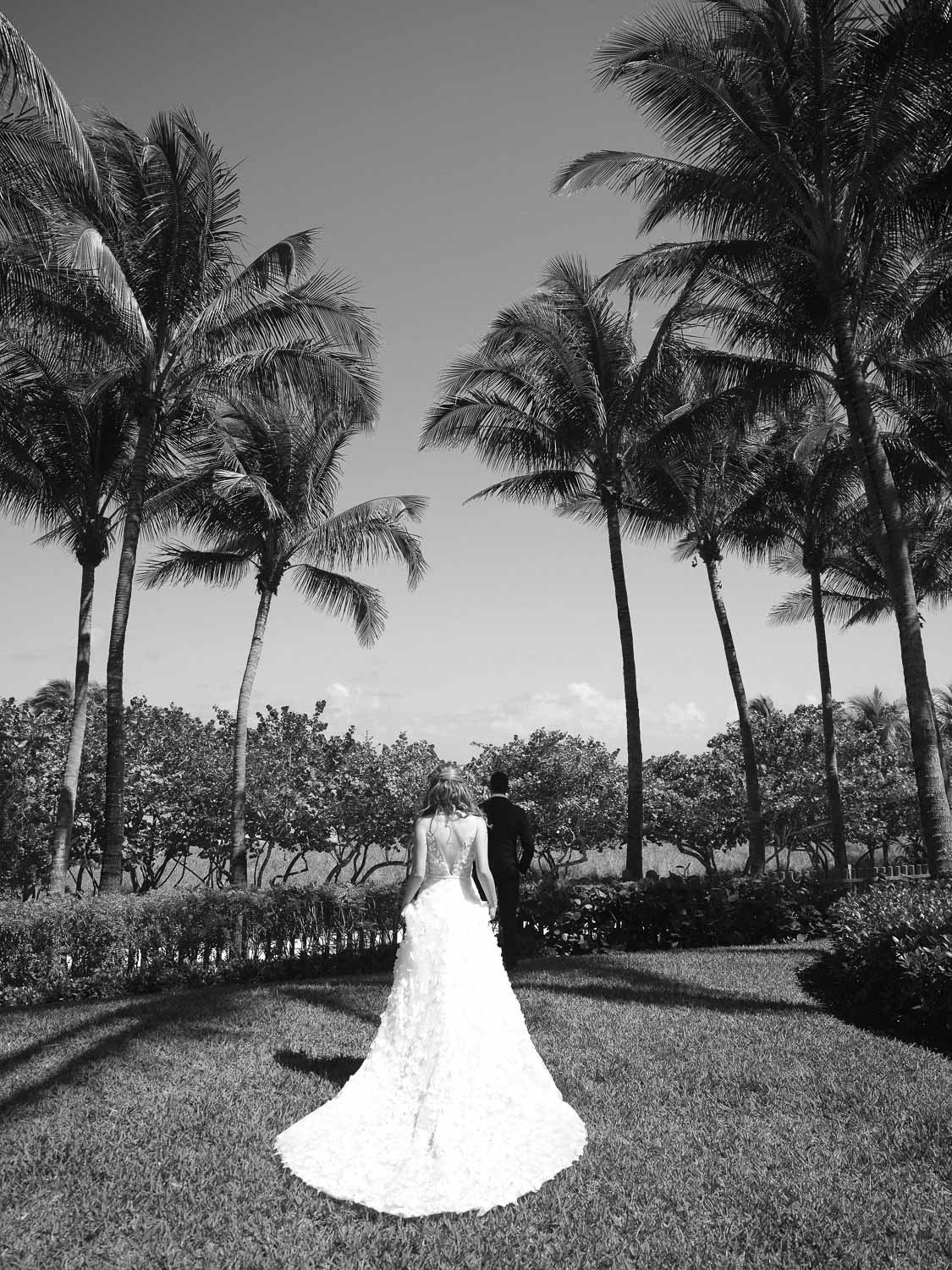 A bride in a wedding dress walking hand-in-hand with a groom in a suit under palm trees in a garden.