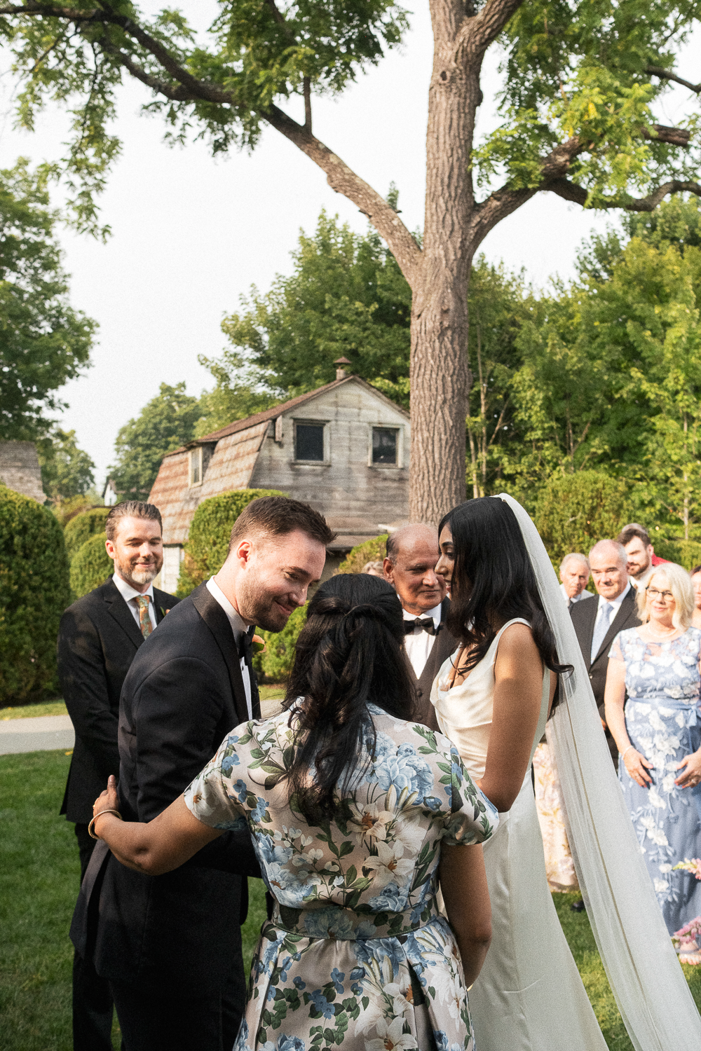 Wedding ceremony outdoors with bride, groom, officiant, and guests under a large tree.