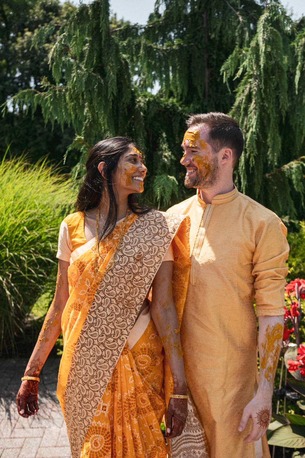 A couple dressed in traditional Indian attire, smiling at each other, with yellow and orange powder on their faces and clothes, outdoors with green trees and plants in the background.