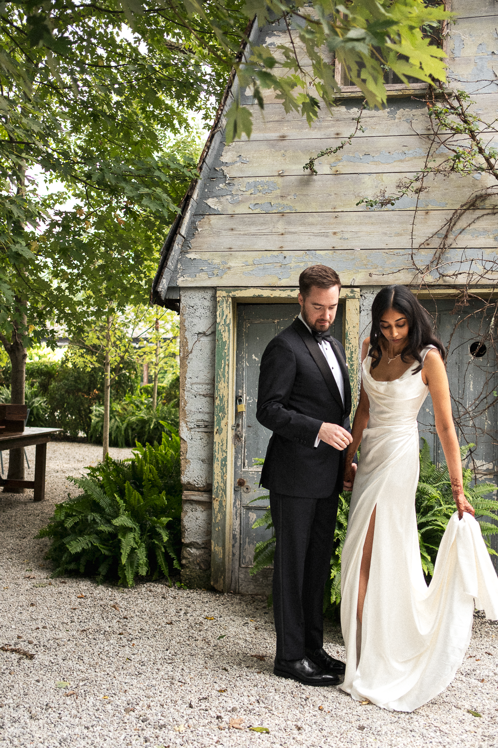 A couple in wedding attire standing outdoors near a weathered wooden shed with greenery and trees around.