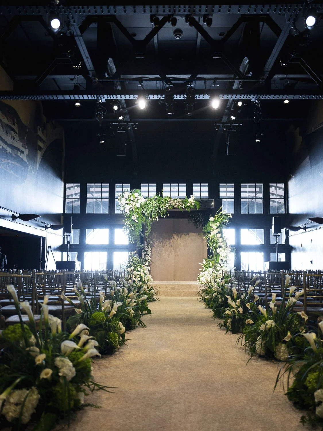 Empty wedding ceremony setup with floral aisle and stage in a large, sunlit hall.