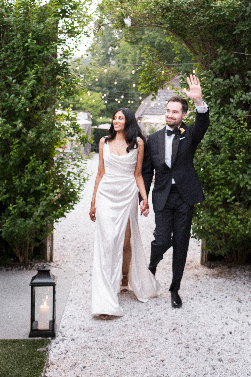 A bride and groom walking hand-in-hand through a garden arch with string lights, bride in a white gown and groom in a tuxedo, waving to guests.