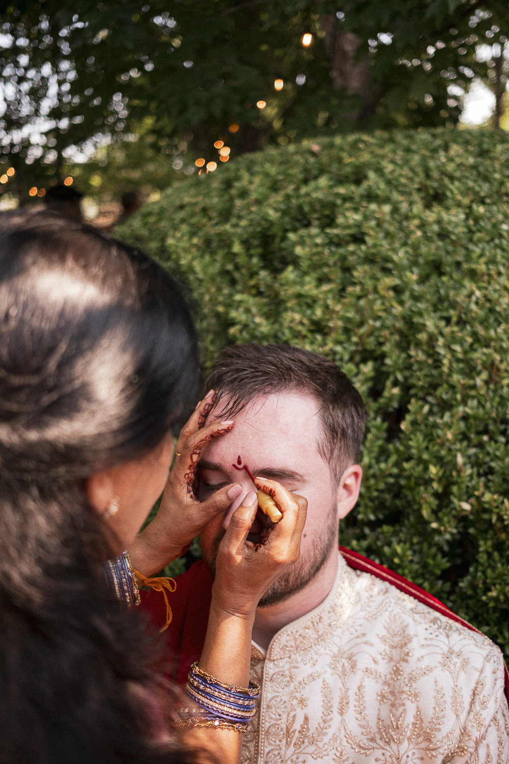 A woman decorating a young man's face with henna at an outdoor event, with greenery and string lights in the background.