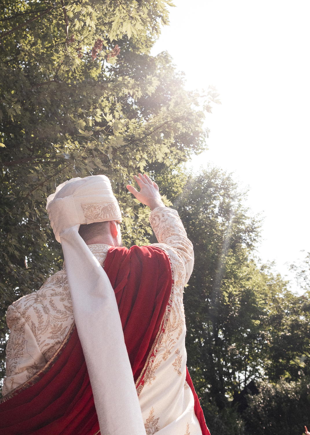 A person dressed in traditional South Asian attire, including a beige embroidered sherwani and a white turban with a beige and gold border, raising their hand towards the sunlight on a clear day with lush green trees in the background.