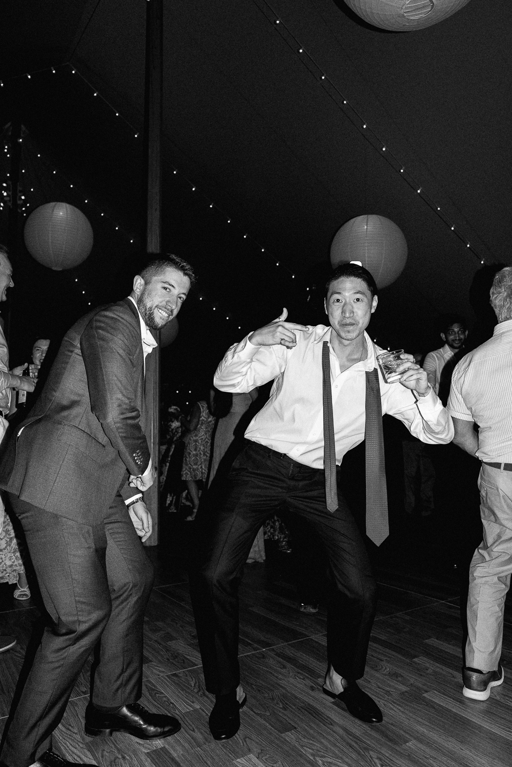 Two men in suits dancing and posing at a celebration or party, with other guests in the background, string lights, paper lanterns, and a dark ceiling.