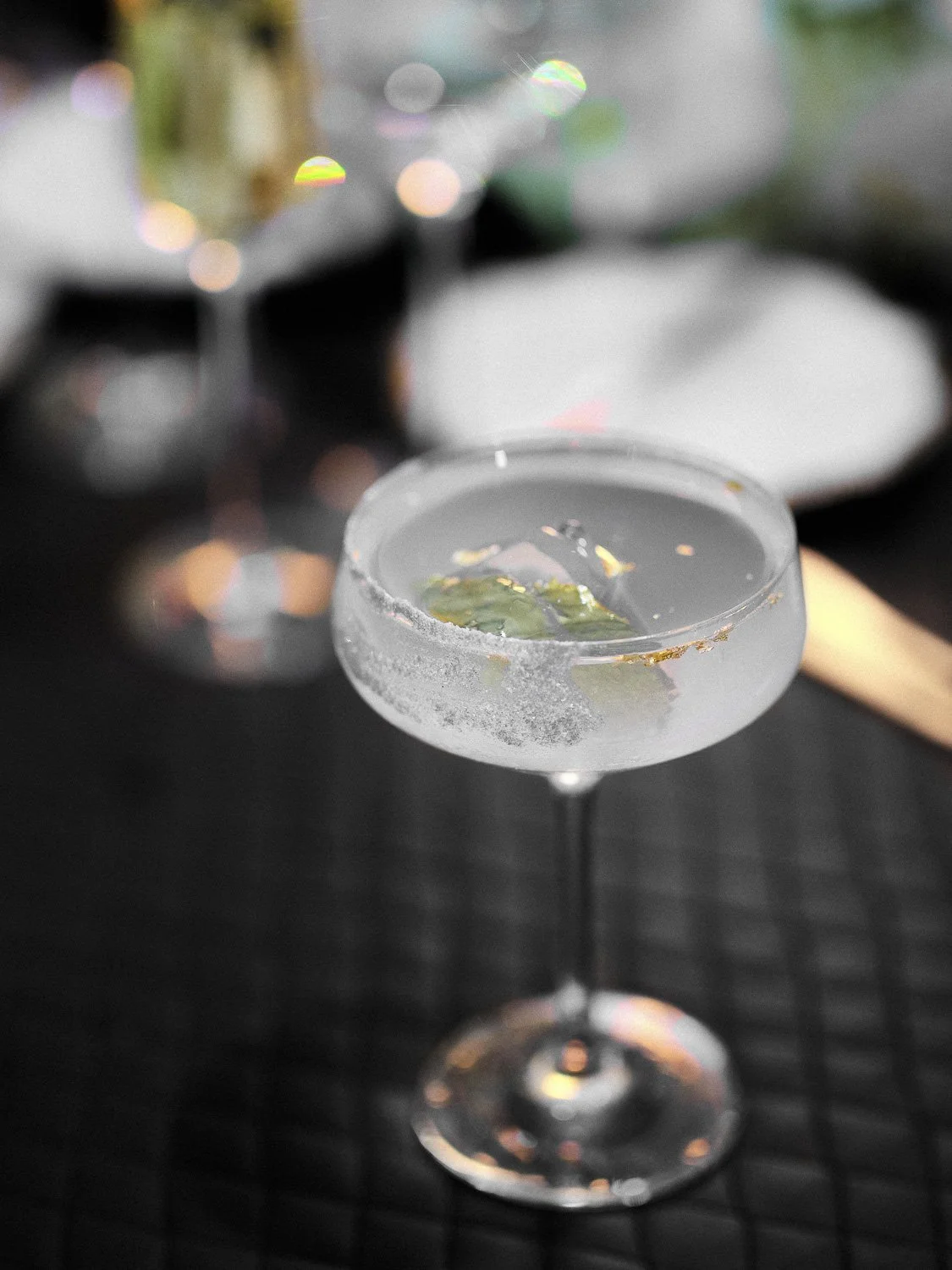 A cocktail glass with a salted rim containing a clear beverage and a lime wedge, set on a black surface with blurred colorful reflections in the background.