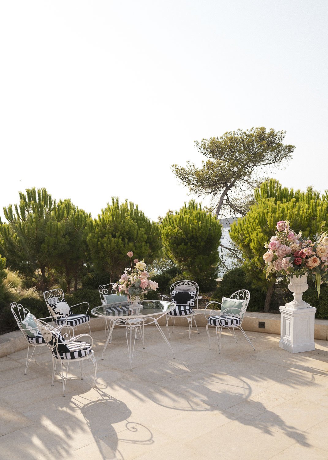 Outdoor patio area with a glass table, six white wrought iron chairs with black and white striped cushions, and large floral arrangements in white urns, surrounded by green trees and shrubs.