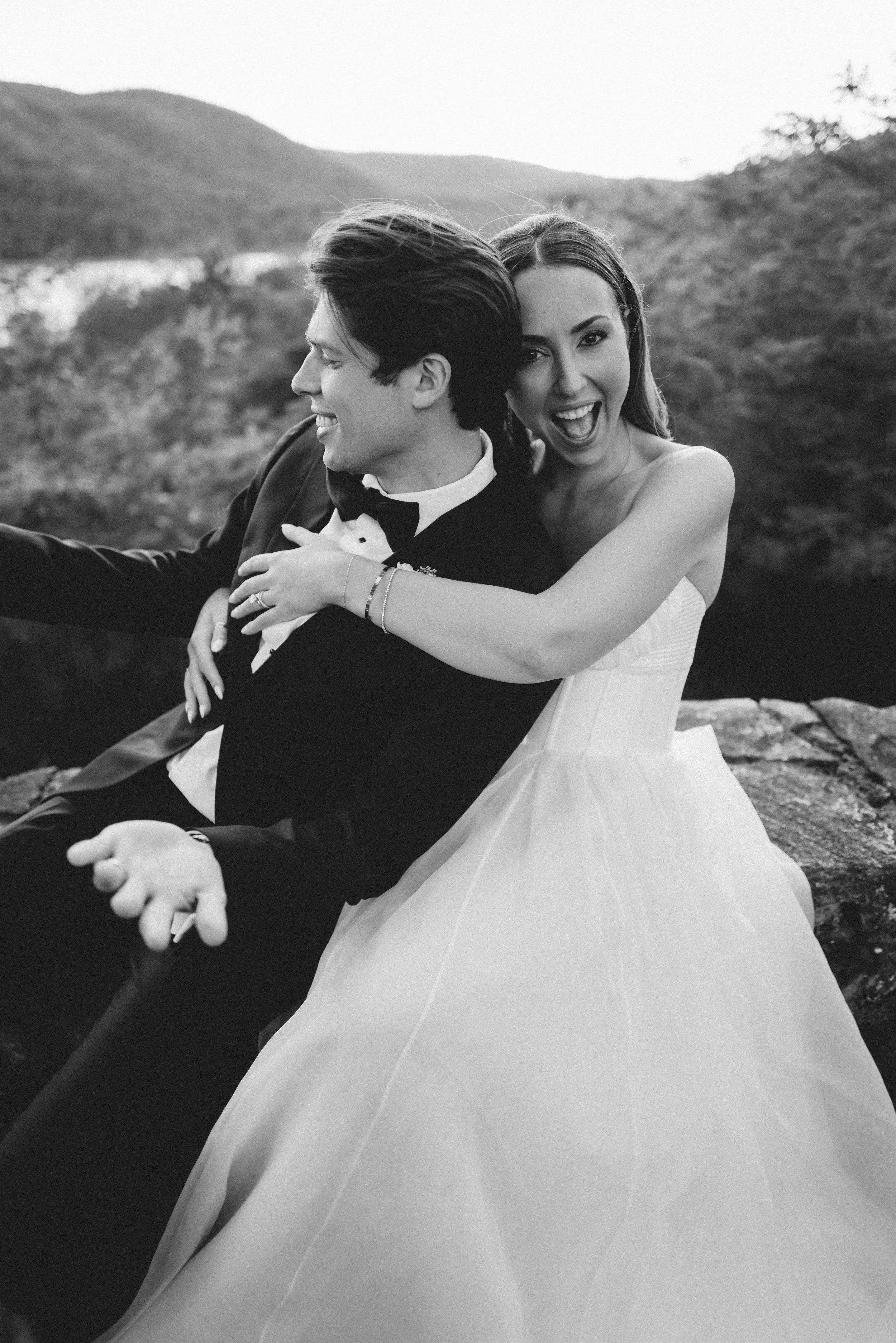 Black and white photo of a bride and groom outdoors. The bride is smiling with her mouth open, hugging the groom from behind. The groom is dressed in a tuxedo and bow tie, smiling with his eyes closed, and the bride's wedding dress has a full skirt. 