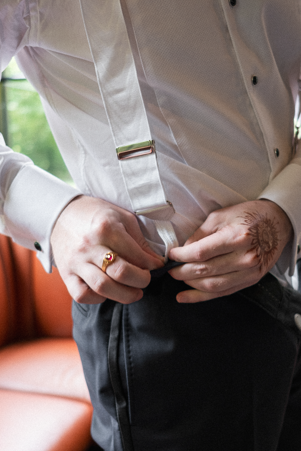 Person buttoning up shirt, wearing a white shirt with suspenders, black pants, and a ring, with a tattoo on one hand, in a room with orange seating and a window in the background.