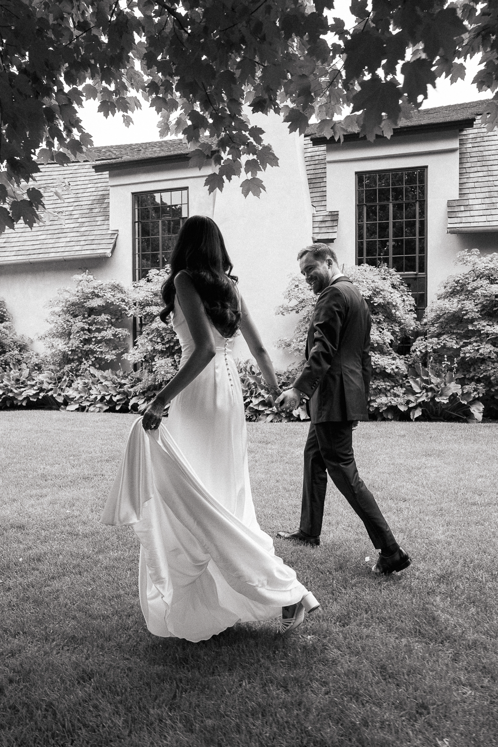 A bride and groom dancing outdoors on a grassy lawn, holding hands, with a house and trees in the background. The bride is wearing a long white gown and the groom a dark suit.