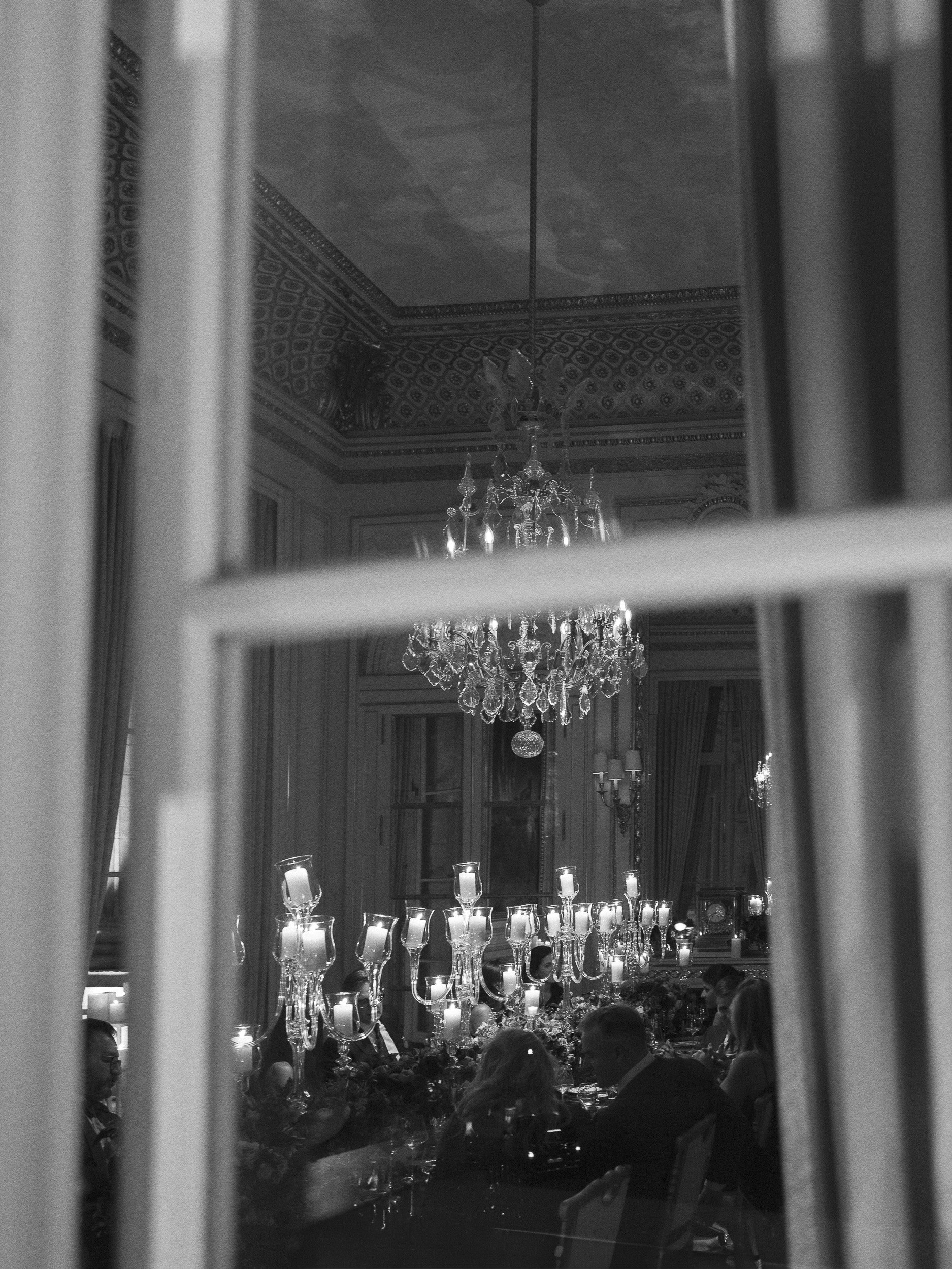 A black and white photo capturing a formal dining room viewed through a window. The room has a chandelier hanging from the ornate ceiling and numerous lit candles on the table below, with several people seated around the table.