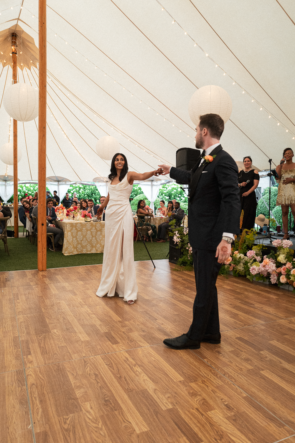 Bride and groom dancing at a wedding reception inside a large white tent with hanging lanterns and string lights, surrounded by seated guests and floral decorations.