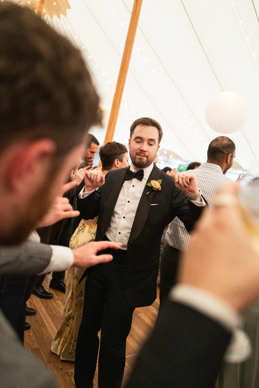 Man in tuxedo and bow tie dancing at a celebration, surrounded by other guests under a white tent with string lights.