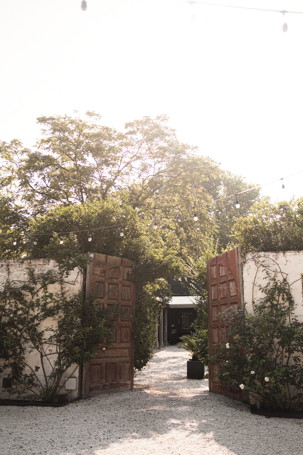 Open wooden gates leading to a gravel pathway with trees and string lights overhead.