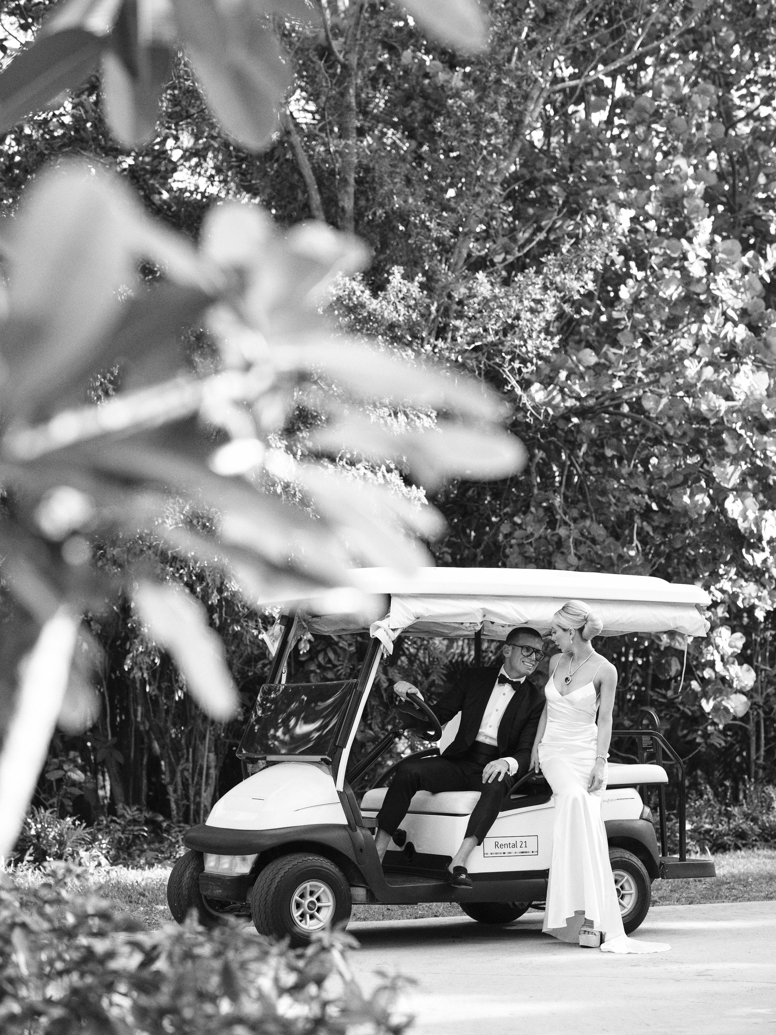 A couple dressed in wedding attire sitting on a golf cart outdoors. The groom is in a tuxedo and the bride is in a white wedding gown, standing beside him, engaged in conversation. The scene is surrounded by trees and foliage.