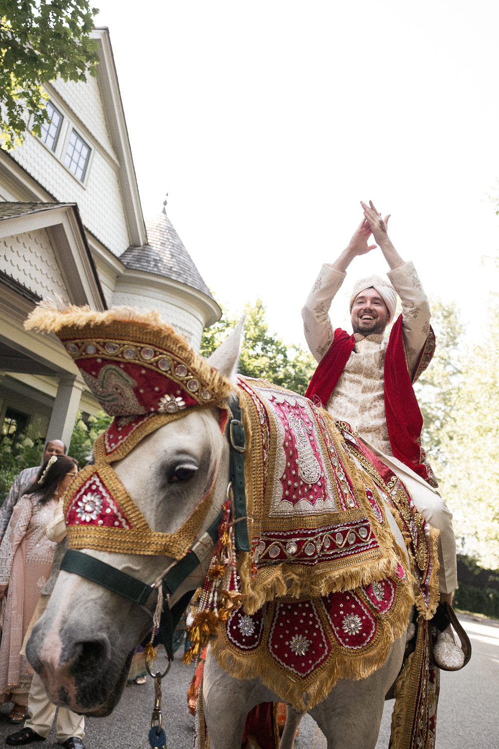 A man in traditional Indian attire riding a decorated white horse during a celebration, with others in festive clothing in the background.