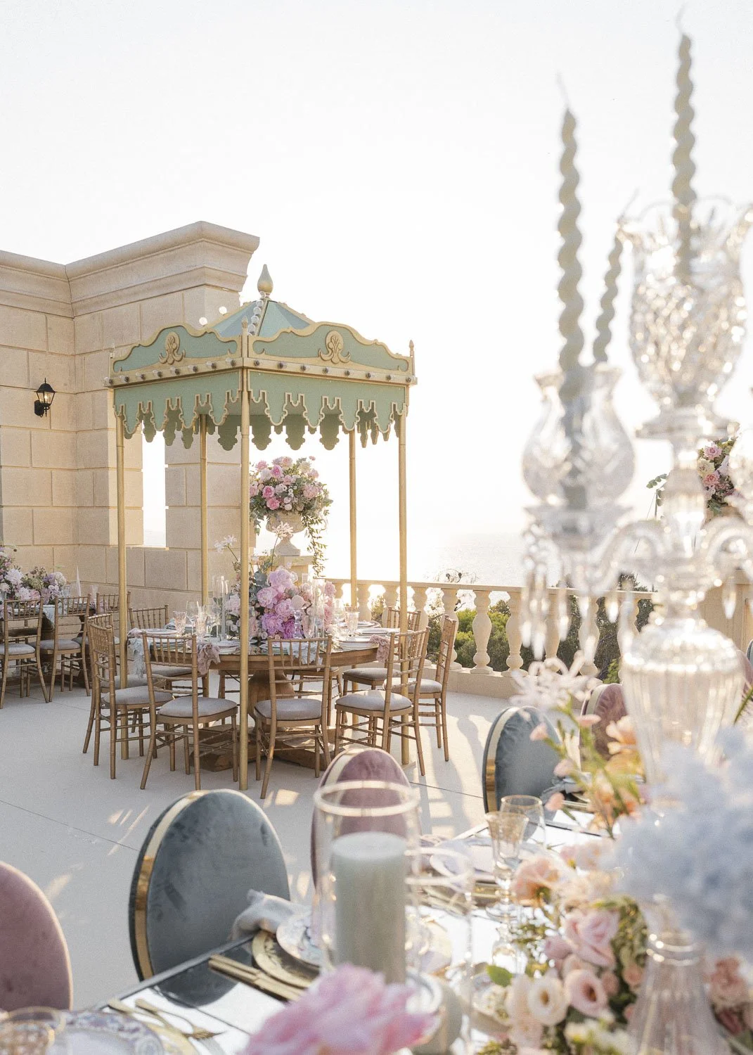 Elegant outdoor dining setup with floral arrangements, wooden chairs, and a vintage-style umbrella on a terrace with a view of the ocean.