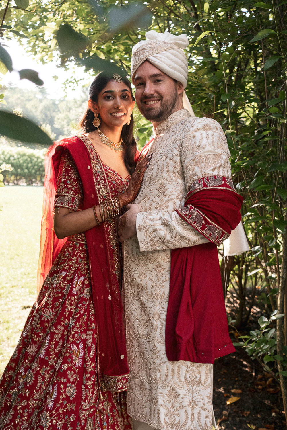 A newlywed couple in traditional Indian wedding attire, standing outdoors among greenery. The bride is wearing a red embroidered lehenga with gold details, jewelry, and henna on her hands. The groom is dressed in a cream and gold embroidered sherwani