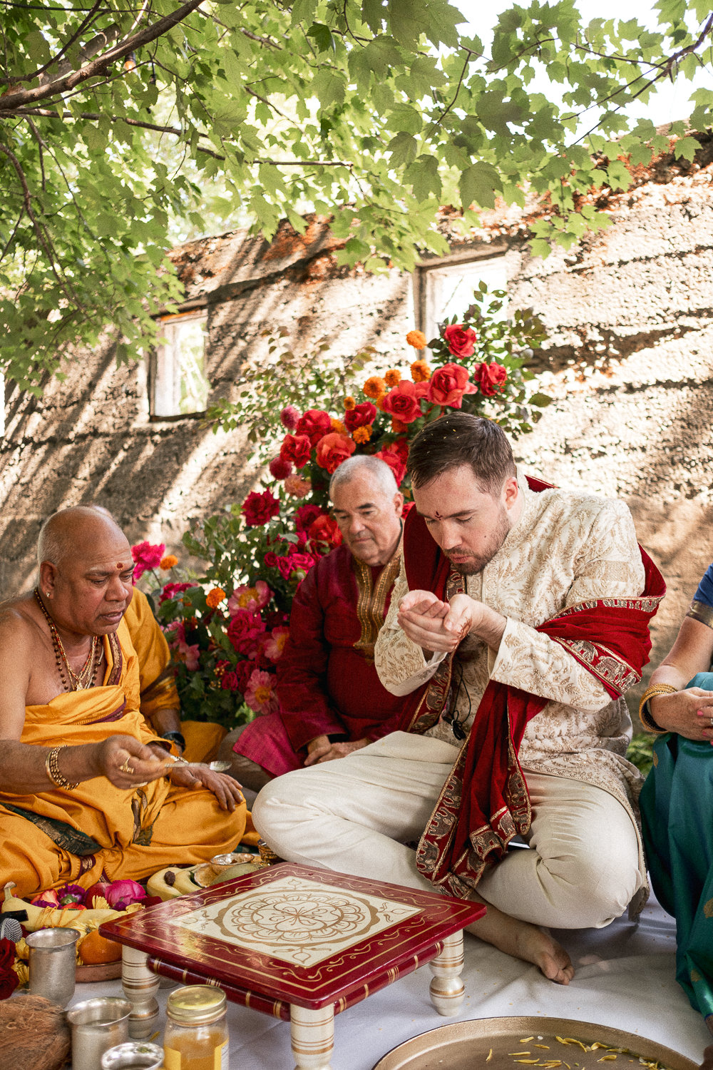 Hindu wedding ceremony with men dressed in traditional attire, sitting on the ground inside a decorated structure with plants and flowers.