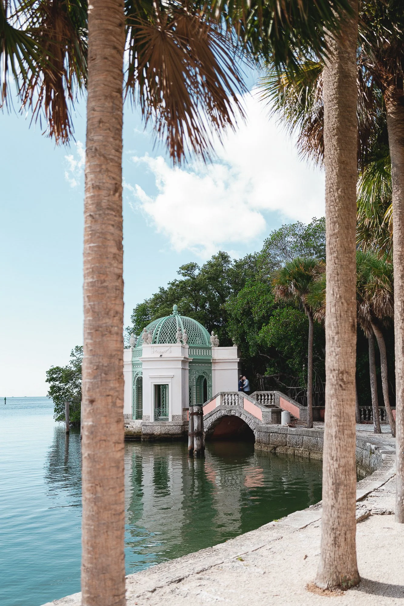 A small decorative pavilion with a green domed roof by a body of water, framed by palm trees, with a walkway and a couple standing near the pavilion, surrounded by lush greenery.