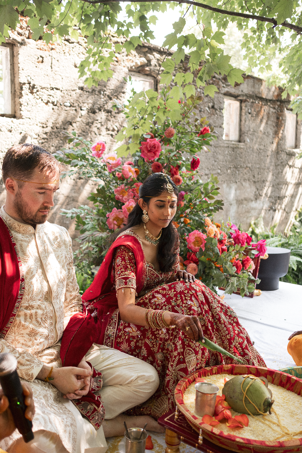 A man and a woman dressed in traditional Indian wedding attire participate in a ceremony outdoors, seated on the ground with flowers and ceremonial objects around them.