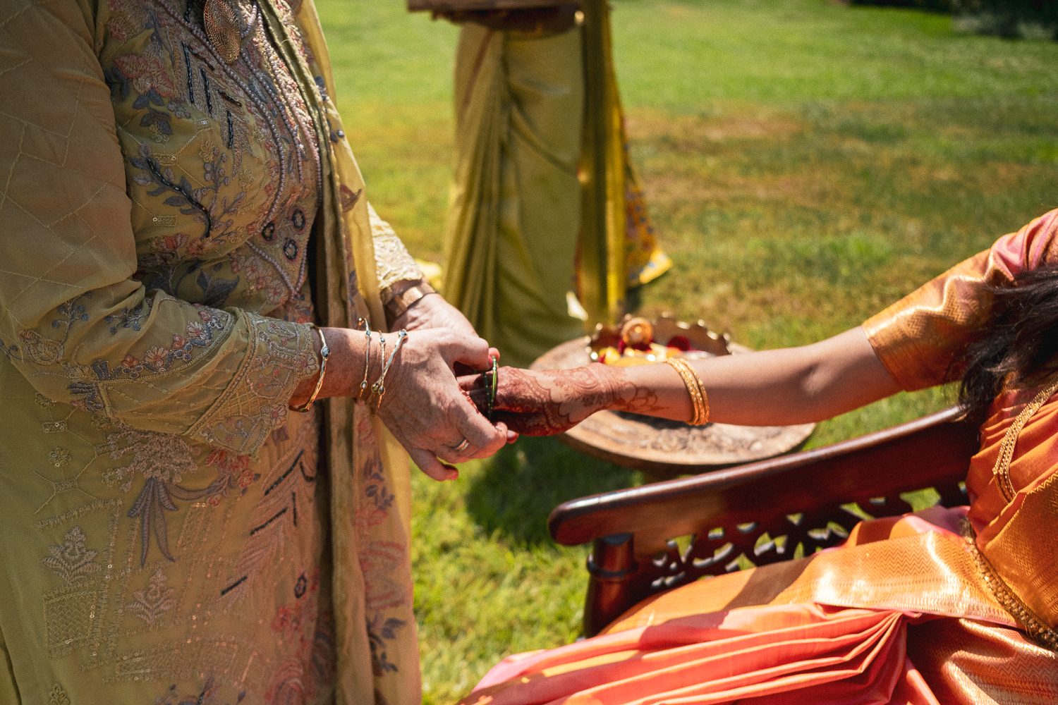 An Indian bride and an older woman holding hands during a traditional ceremony outdoors. The bride is dressed in orange saree with gold accents, and the older woman is wearing a yellow embroidered garment, both adorned with jewelry.