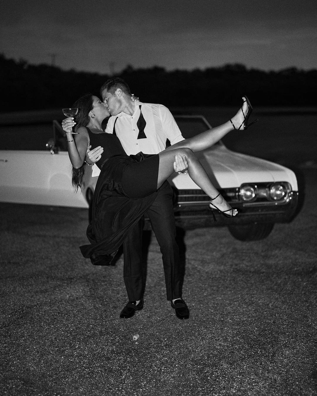 A man in a tuxedo holding a woman in a black dress in his arms while they kiss in front of a vintage car during nighttime.