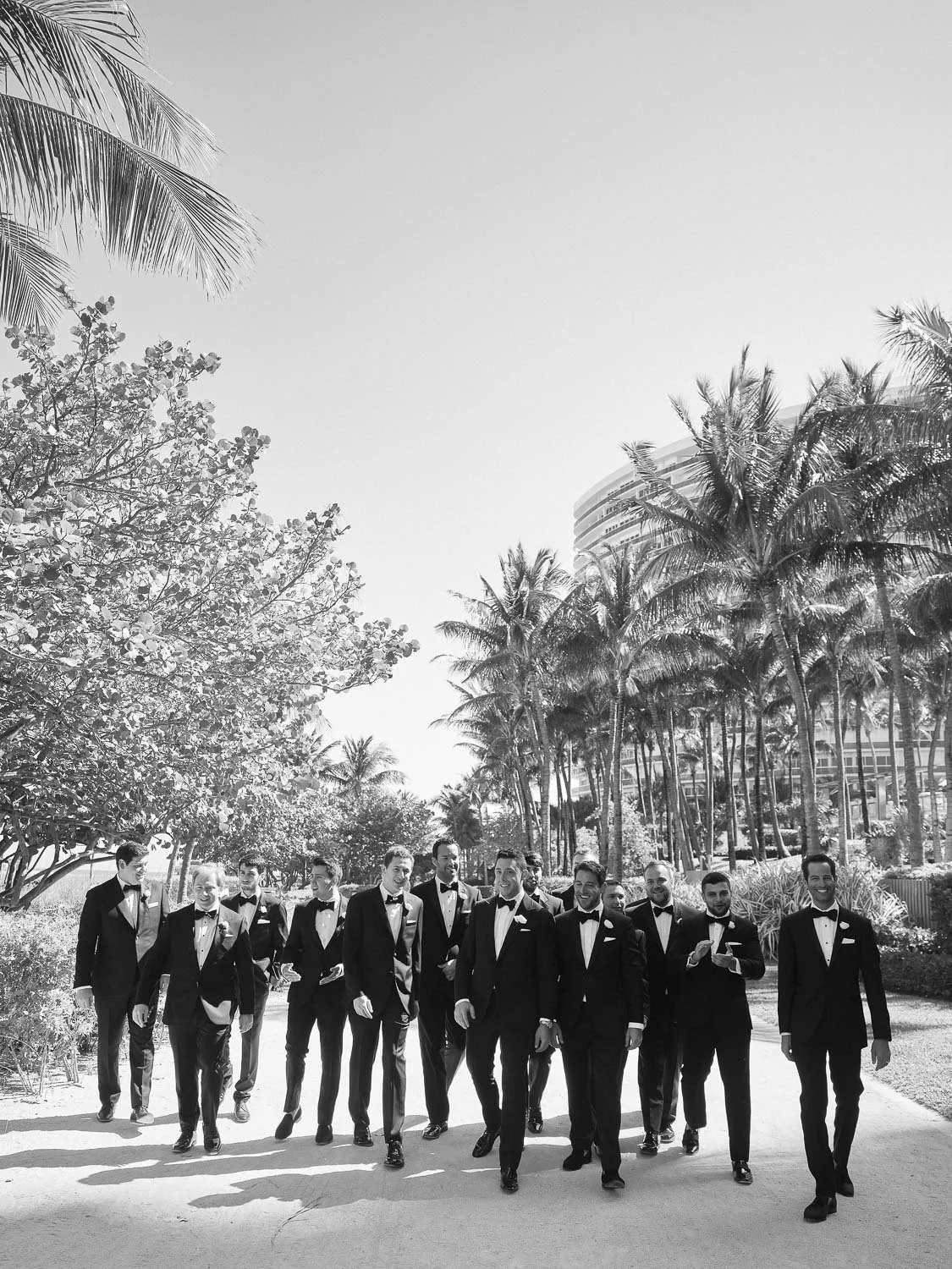 Group of men in tuxedos walking outdoors among palm trees and modern buildings.