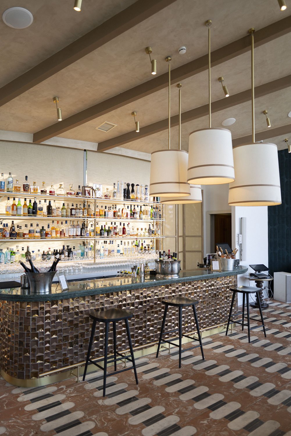 Luxury bar with a curved, reflective counter, three large white pendant lights, and a back wall with shelves stocked with various bottles of alcohol.