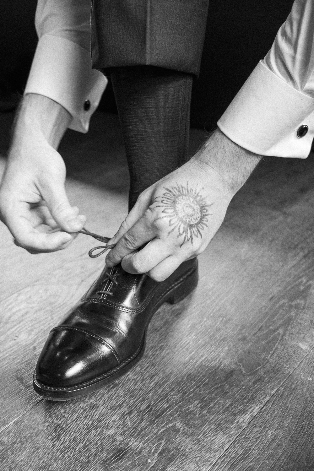 A person wearing dress shoes and dark pants is getting their shoelace tied, with hands visible and one hand having a sun tattoo.