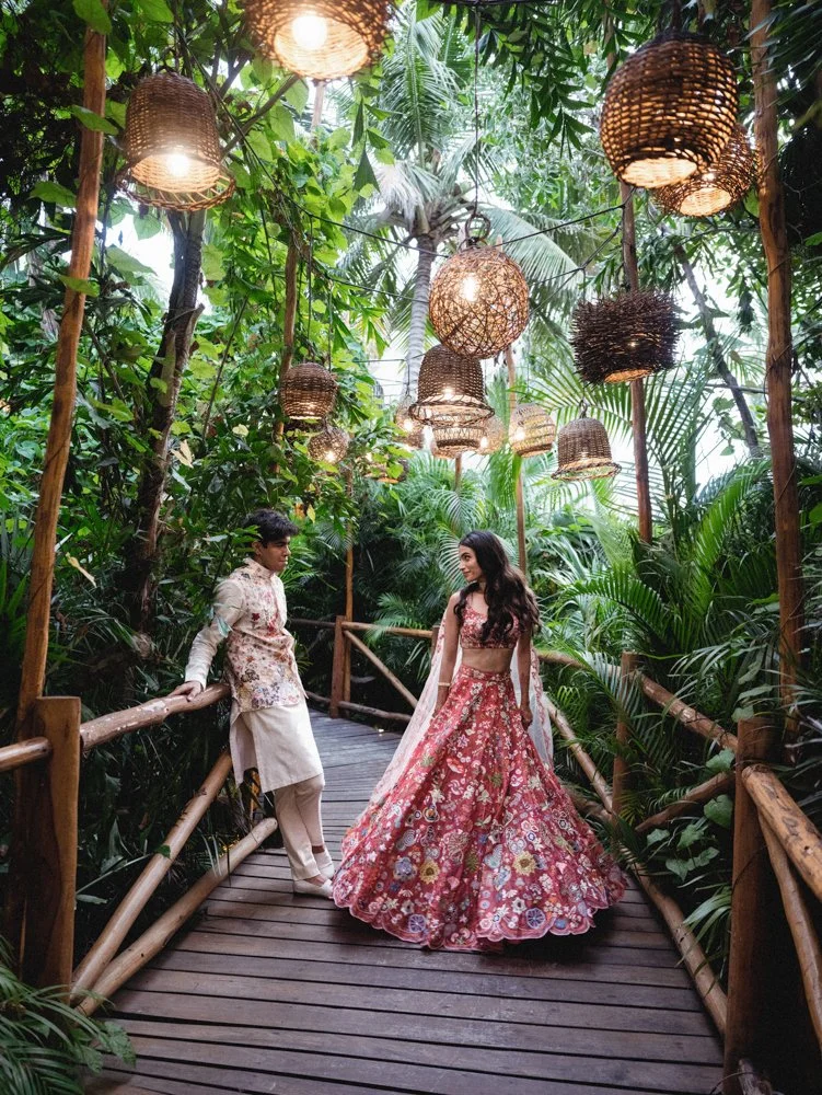A couple dressed in traditional South Asian attire standing on a wooden pathway in a lush, tropical forest decorated with hanging woven lanterns.