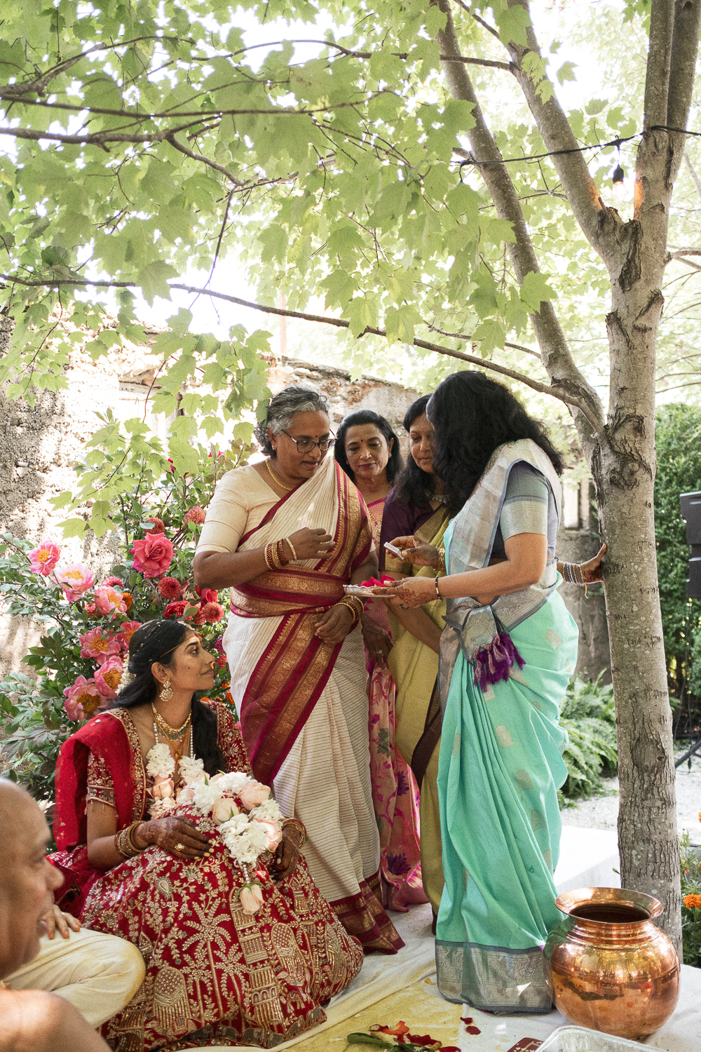 Women in traditional Indian sarees participating in a cultural ceremony outdoors under a tree, with flowers and pottery around.