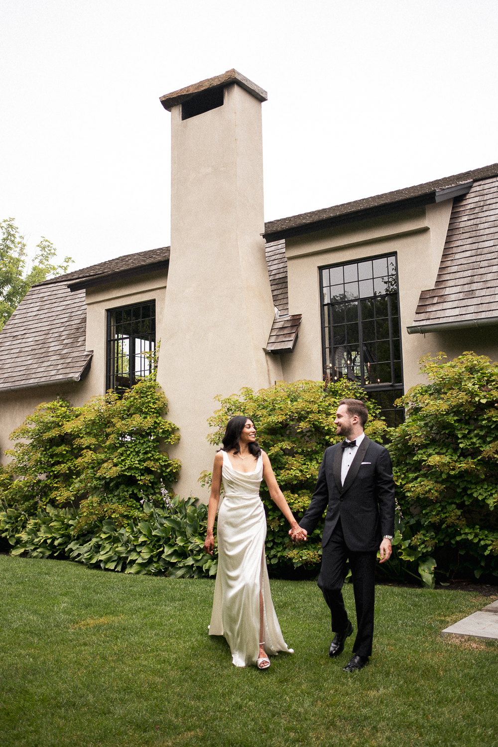 A couple in wedding attire walking hand in hand on a lawn, with a house and lush greenery in the background.