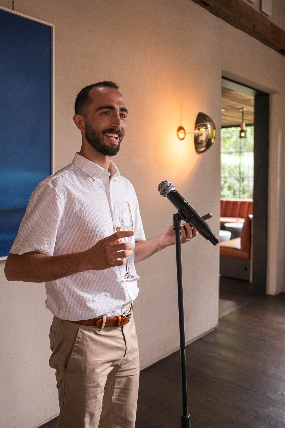 A man in a white shirt and khaki pants holding a glass of champagne and speaking at a microphone in a warmly lit room.