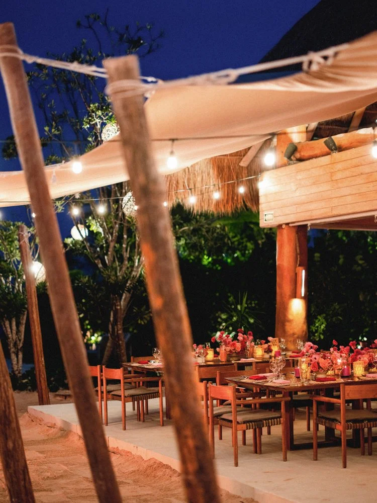 Outdoor dining area decorated with pink flowers, candles, and string lights at night, with wooden tables and chairs surrounded by trees.
