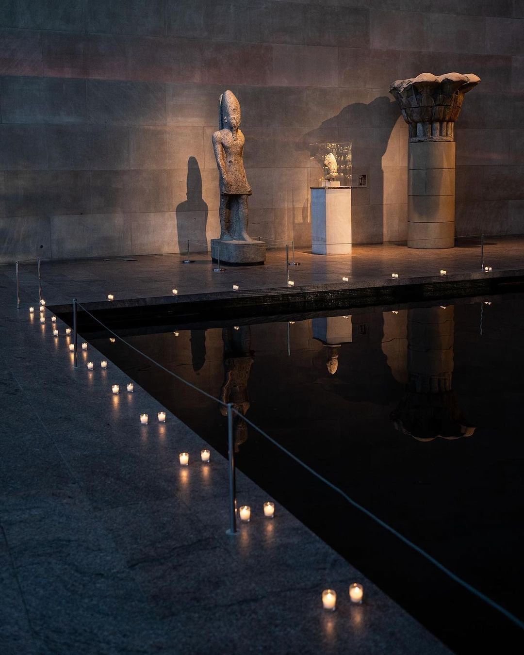 Ancient sculptures displayed in a modern museum with candles and a reflective pool of water.
