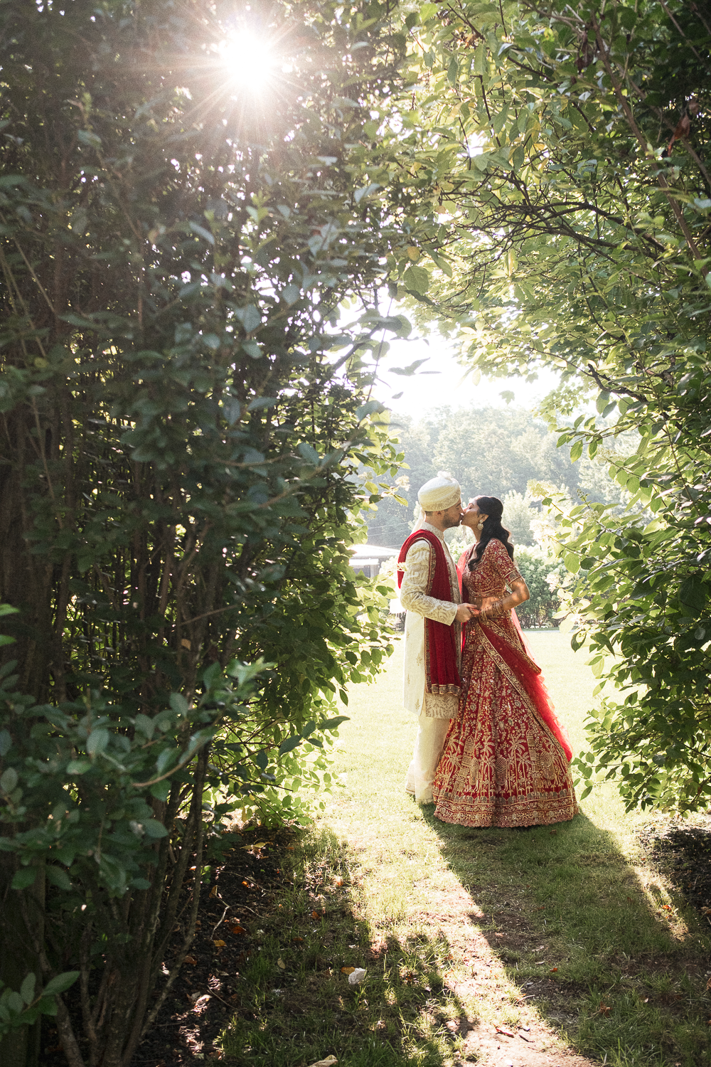 A couple dressed in traditional Indian wedding attire kissing in a sunlit garden, framed by dense greenery.