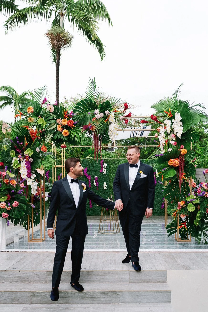 Two men in tuxedos holding hands at a wedding ceremony with a tropical floral arch and palm trees in the background.