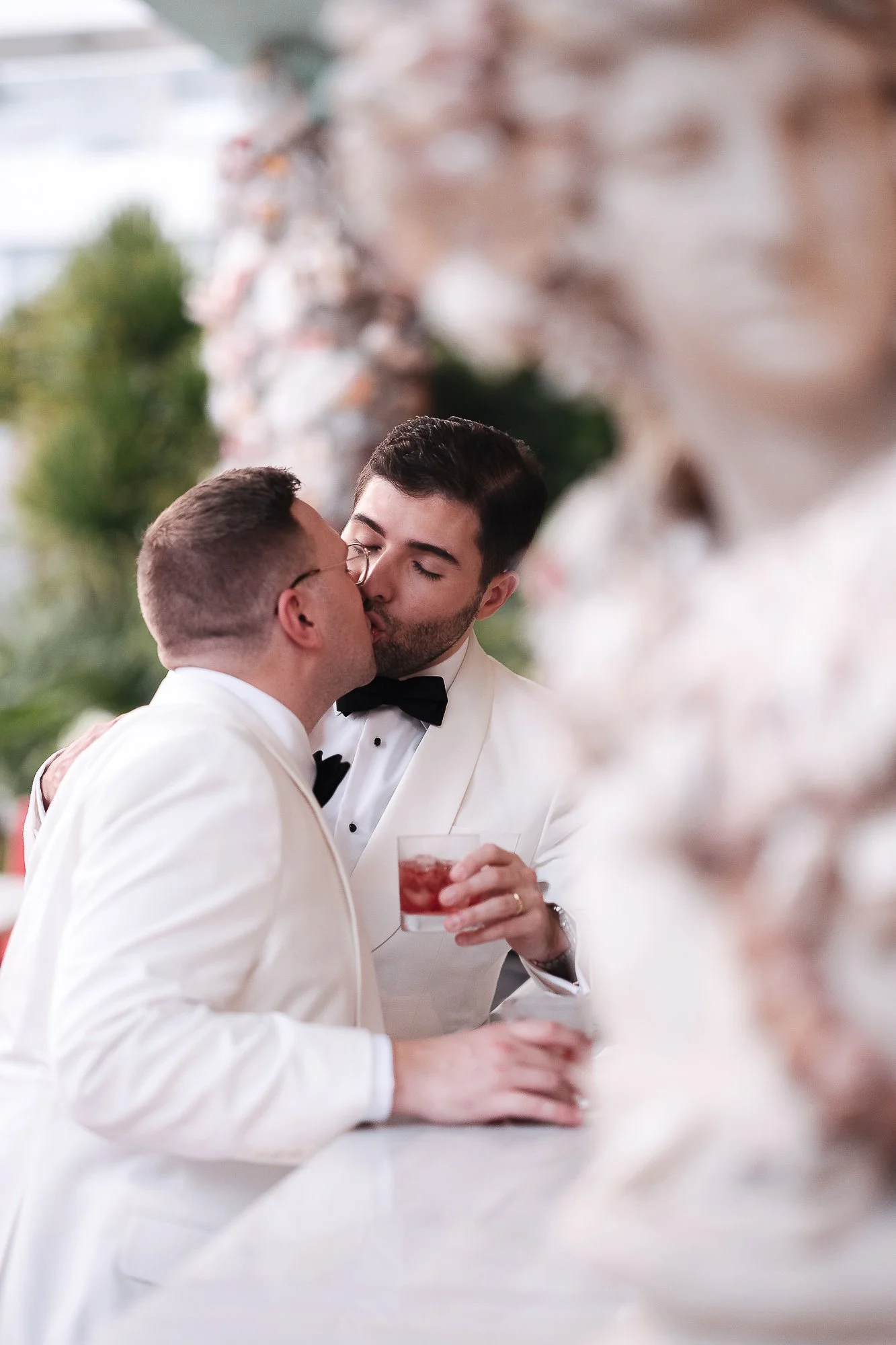 Two men in white tuxedos sharing a kiss at a wedding reception, one holding a glass of red drink, blurred floral decorations in the foreground.