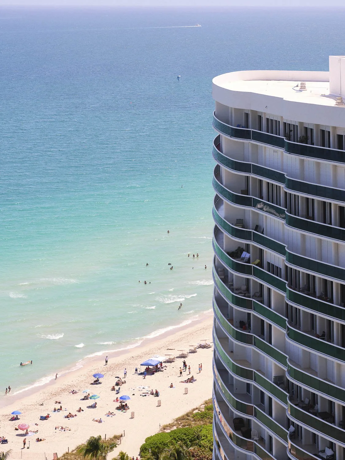 Tall beachfront hotel with curved balconies overlooking a sandy beach and turquoise ocean, with people sunbathing, swimming, and parasailing.
