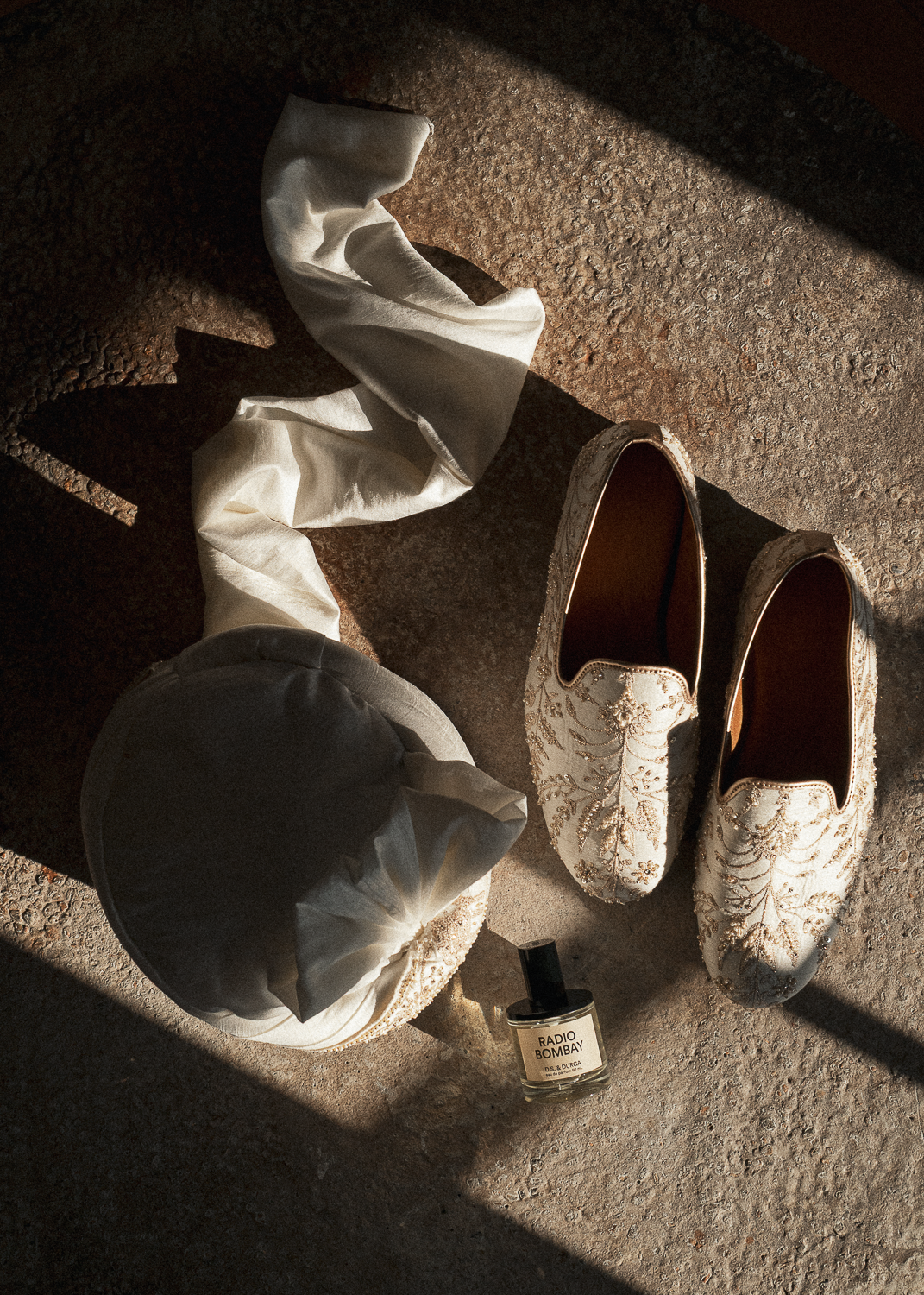 A pair of ornate white embroidered slippers, a crumpled white cloth, a rolled white cloth, a bottle of Radio Bombay perfume, and a beige beret on a textured stone surface with sunlight casting shadows.
