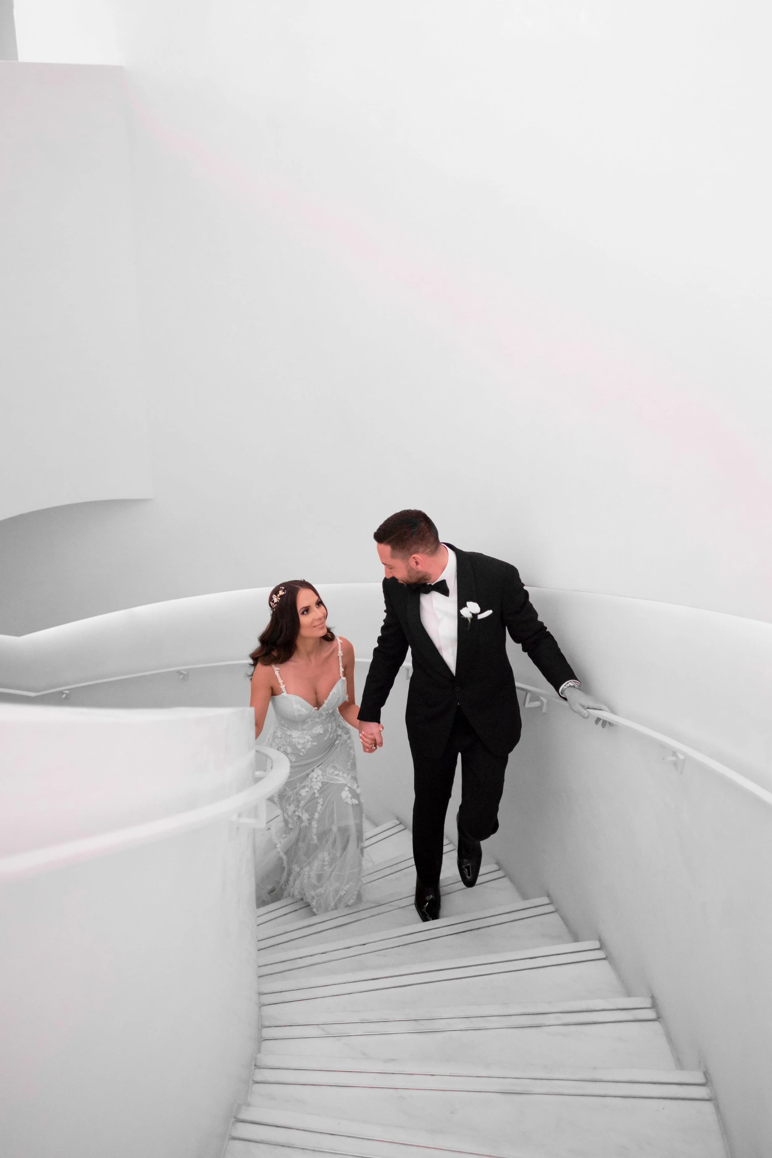A bride and groom holding hands on a white spiral staircase, with the bride in a white wedding dress and the groom in a tuxedo, in a modern white interior.