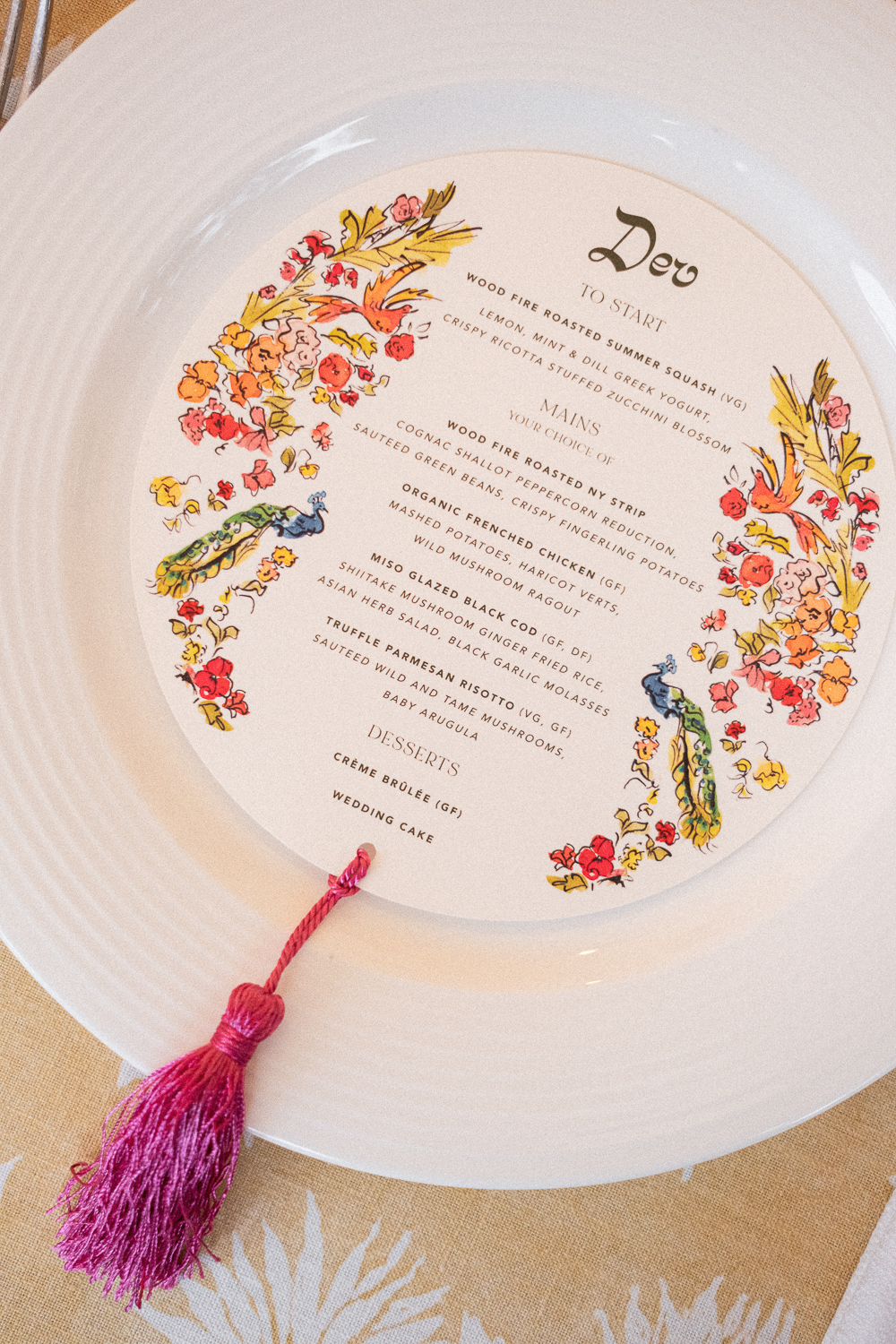 A menu card with floral decorations and a pink tassel attached, featuring a dinner menu for an event called 'Deo' with sections for starters, mains, and desserts.