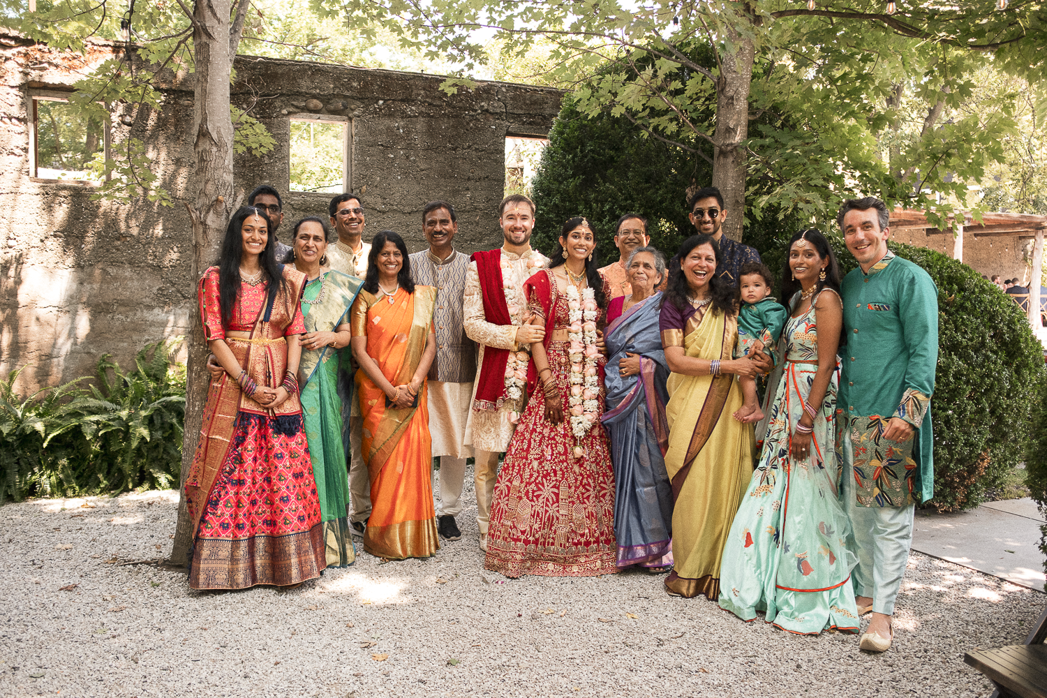 A large group of people in traditional Indian clothing posing outdoors for a wedding celebration, with trees and a stone building in the background.