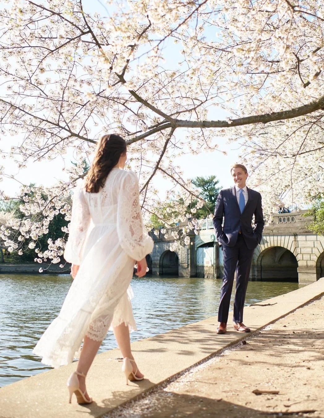 A woman in a white lace dress and heels walking along a riverbank, approaching a man in a navy suit and blue tie who is standing with his hands in his pockets. They are beneath blooming cherry blossom trees near a stone bridge.
